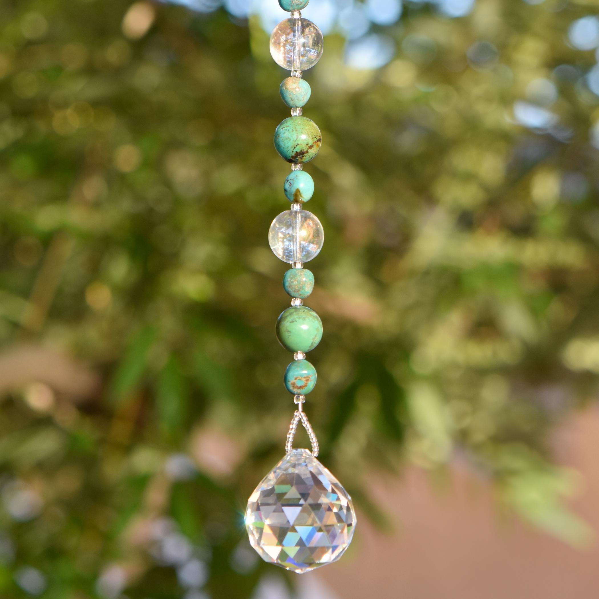 May include: A hanging crystal suncatcher with a string of turquoise and clear beads. The faceted crystal at the bottom reflects light, creating a sparkling effect. The beads are strung on a silver-colored wire.