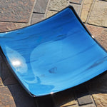 Six inch square glass tray with blue and black streaked center, black trim and black base, resting on stone tile table in sunlight.