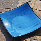 Six inch square glass tray with blue and black streaked center, black trim and black base, resting on stone tile table in sunlight.