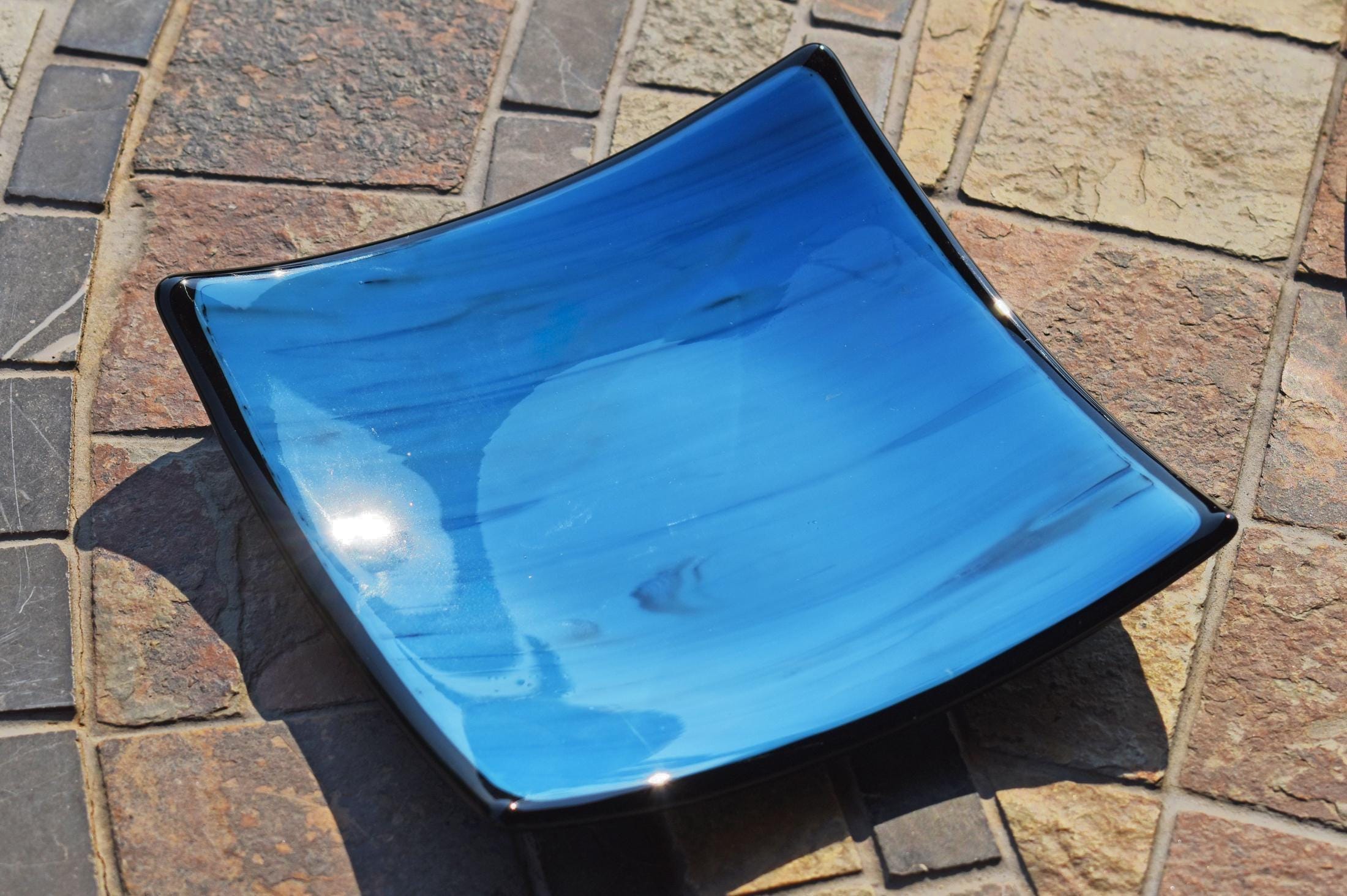 Six inch square glass tray with blue and black streaked center, black trim and black base, resting on stone tile table in sunlight.