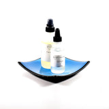 Square fused glass dish with solid blue center, black trim and black base, sitting white surface. Two bottles of skin care products are placed in the tray showing how it will work as a bathroom tray.