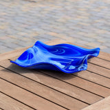 Fused glass square bowl made with medium blue and white swirled glass. The edges of the bowl are wavy and the bowl has a short foot at its center. It is resting on a wooden slat table with brick pavers in the background.
