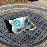 Southwestern-style fused glass bowl on stone and metal table. Bowl is 10" square with wave edges and a small foot. The glass has a swirled pattern of blues, oranges, grays and greens.