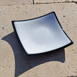 Six inch white glass square tray on stone tile in sunlight. Glass is opaque white with black edges and base.