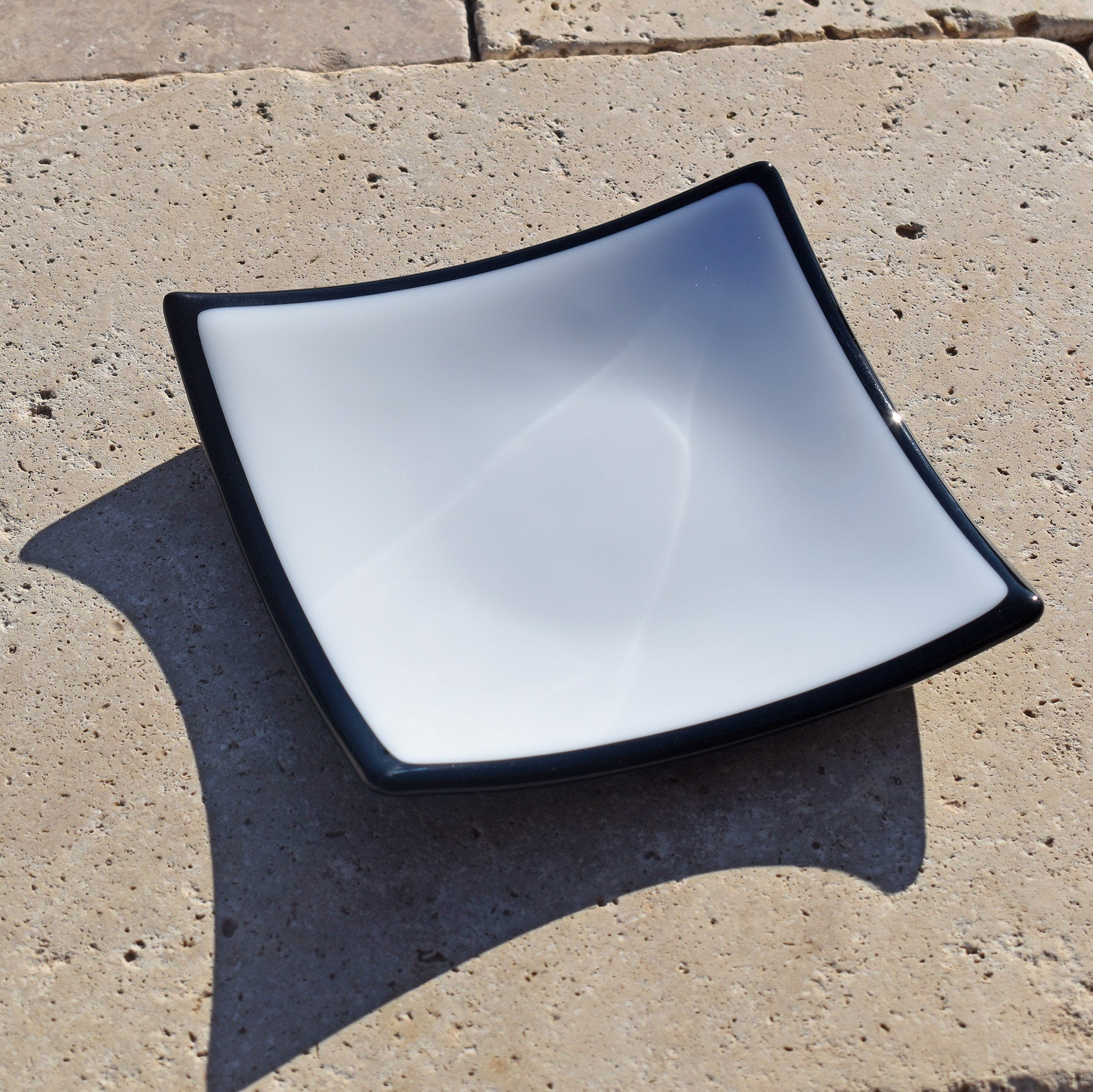 Six inch white glass square tray on stone tile in sunlight. Glass is opaque white with black edges and base.