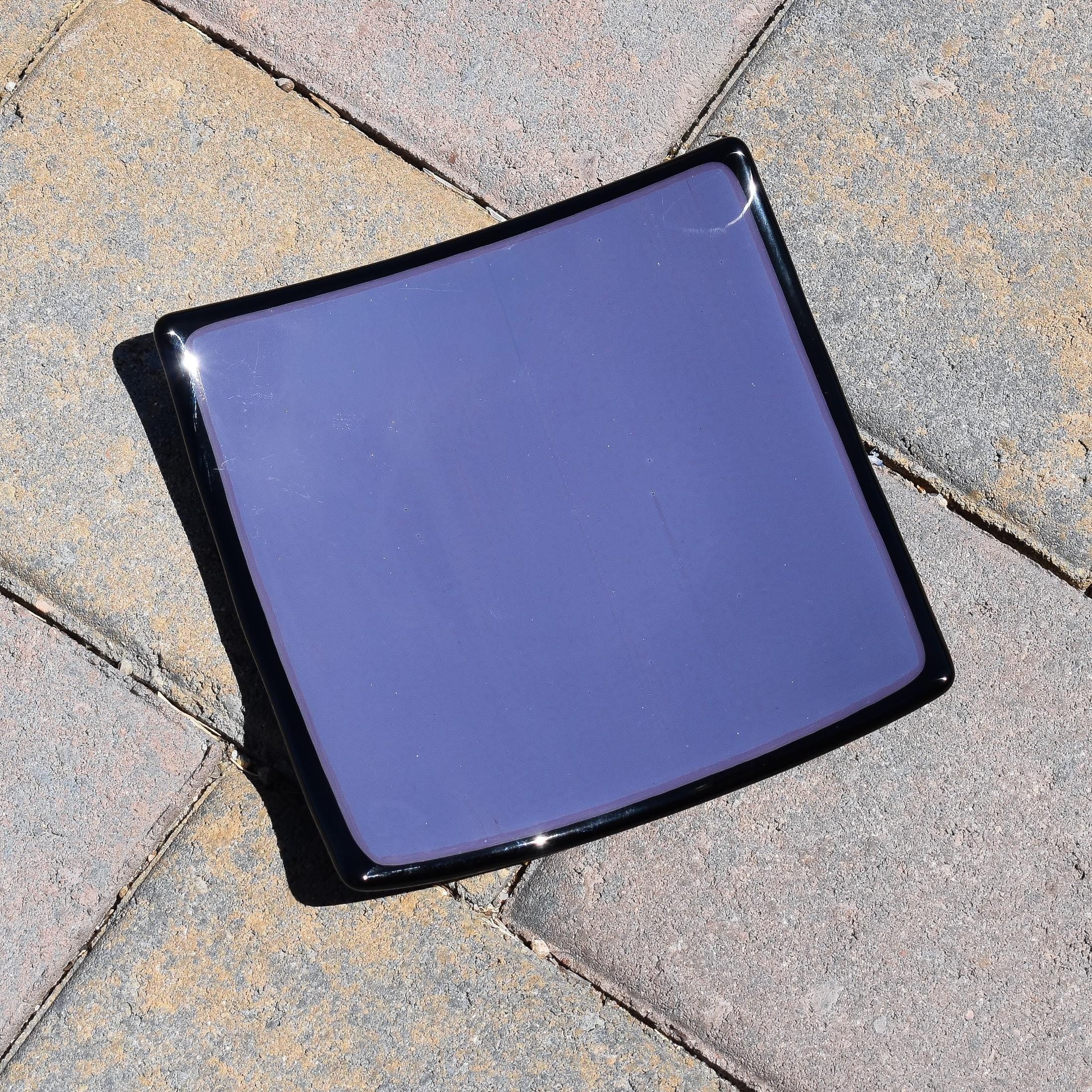 Dusty Violet Purple fused glass tray with black trim sitting on pavers in sunlight. Tray is 6" square.