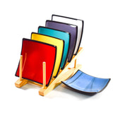 Six different colors of 6 inch square fused glass trays. Five are in a wooden stand and the six, a blue tray is to the side. White Background.