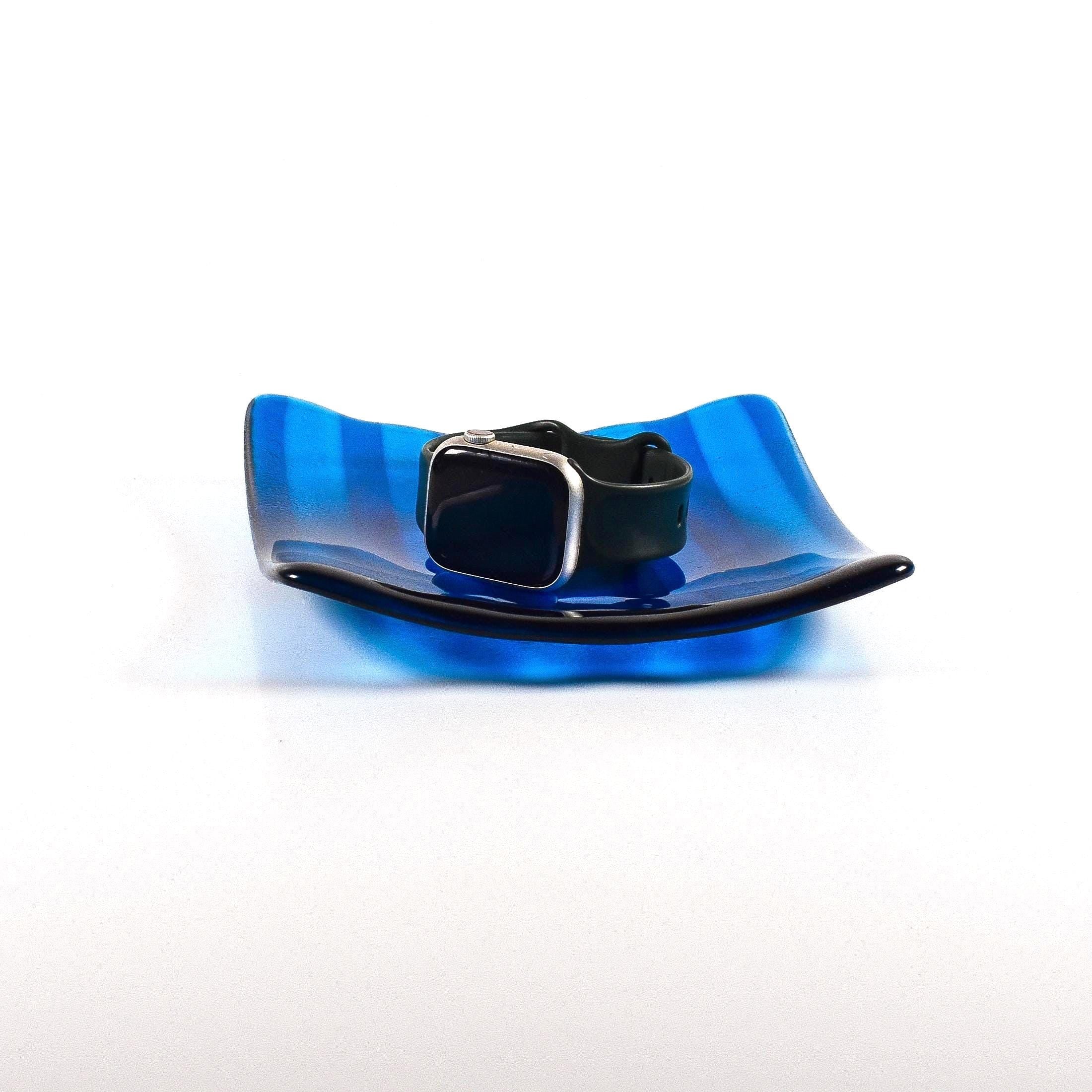 Deep aqua blue fused glass tray with two opposite scalloped edges and strips of a lighter color. The photo is looking at the front of the tray and the tray is on a white surface. The tray is holding a watch to show size and use.