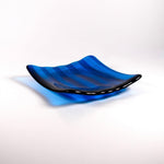 Deep aqua blue fused glass tray with two opposite scalloped edges and strips of a lighter color. The tray is sitting at an angle on a white surface.