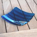 Deep aqua blue fused glass tray with two scalloped edges and strips of a lighter color. The tray is sitting on a wood table at an angle, outdoors, with some reflections in the glass.