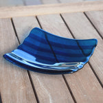Deep aqua blue fused glass tray with two scalloped edges and strips of a lighter color. The tray is sitting on a wood table at an angle, outdoors, with some reflections in the glass.