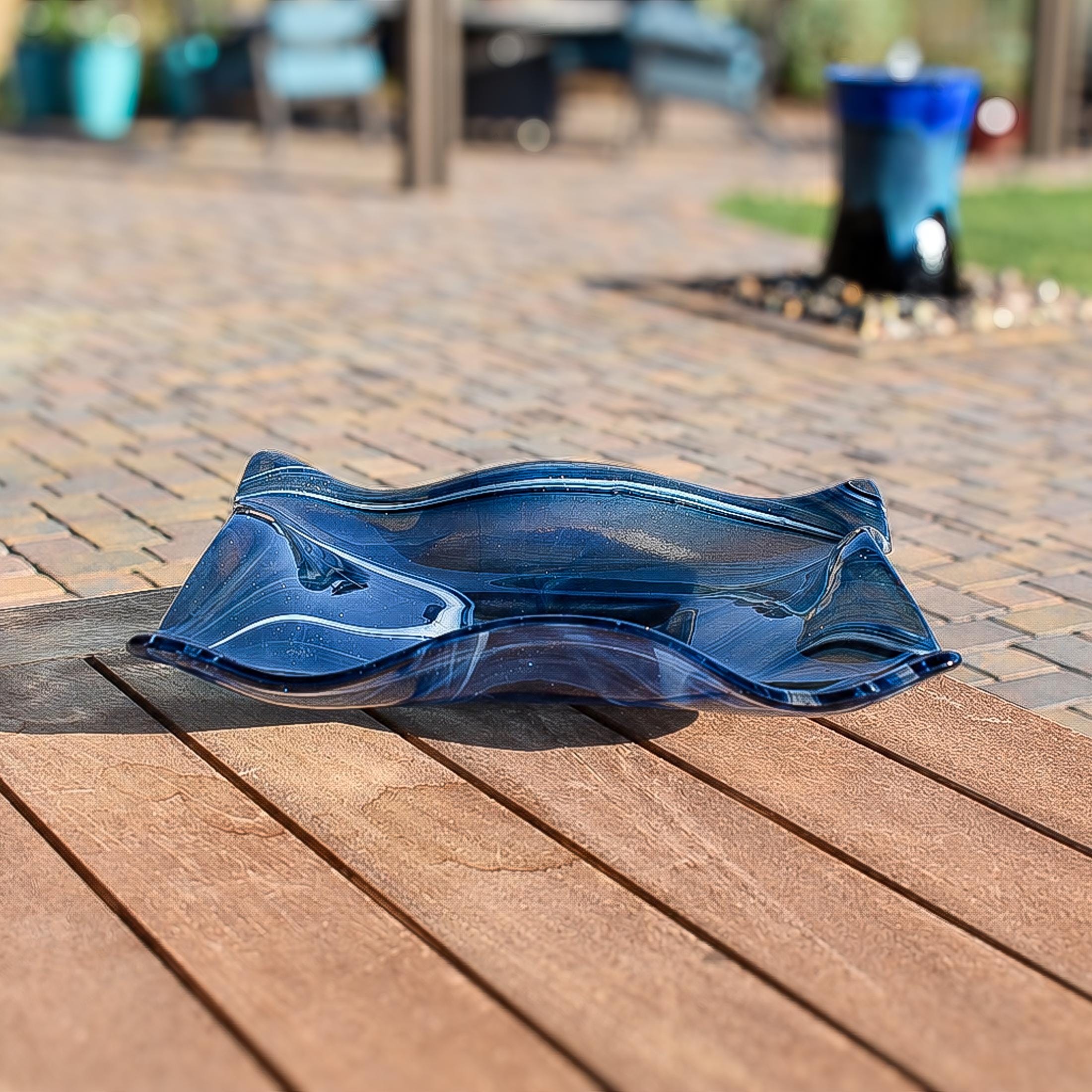 Square, wave edged fused glass bowl sitting on slatted wooden table outdoors. Bowl is 10" square made of transparent aventurine blue glass with swirls of white and a hint of lavender. The transparent aventurine glass sparkles, making the bowl look like it has glitter.