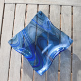 Blue fused glass wave bowl with flowing lines in transparent blue fused glass, shown empty on a slatted wooden table.