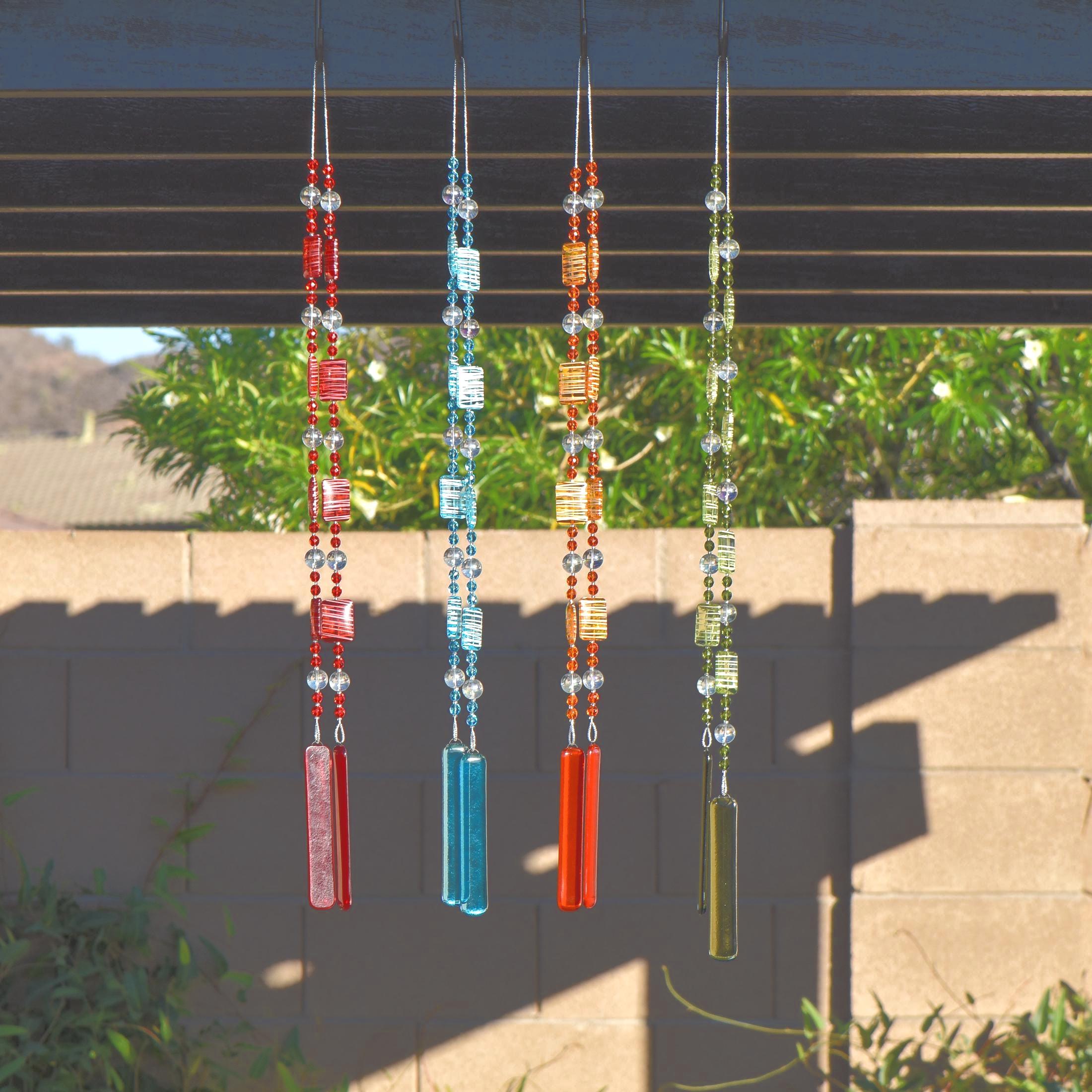 Four glass and acrylic wind chimes hanging from a brown pergola in the sun. Each chime is made with fused glass, glass beads and rectangular acrylic beads. Available in Red, Orange, Green and Blue.