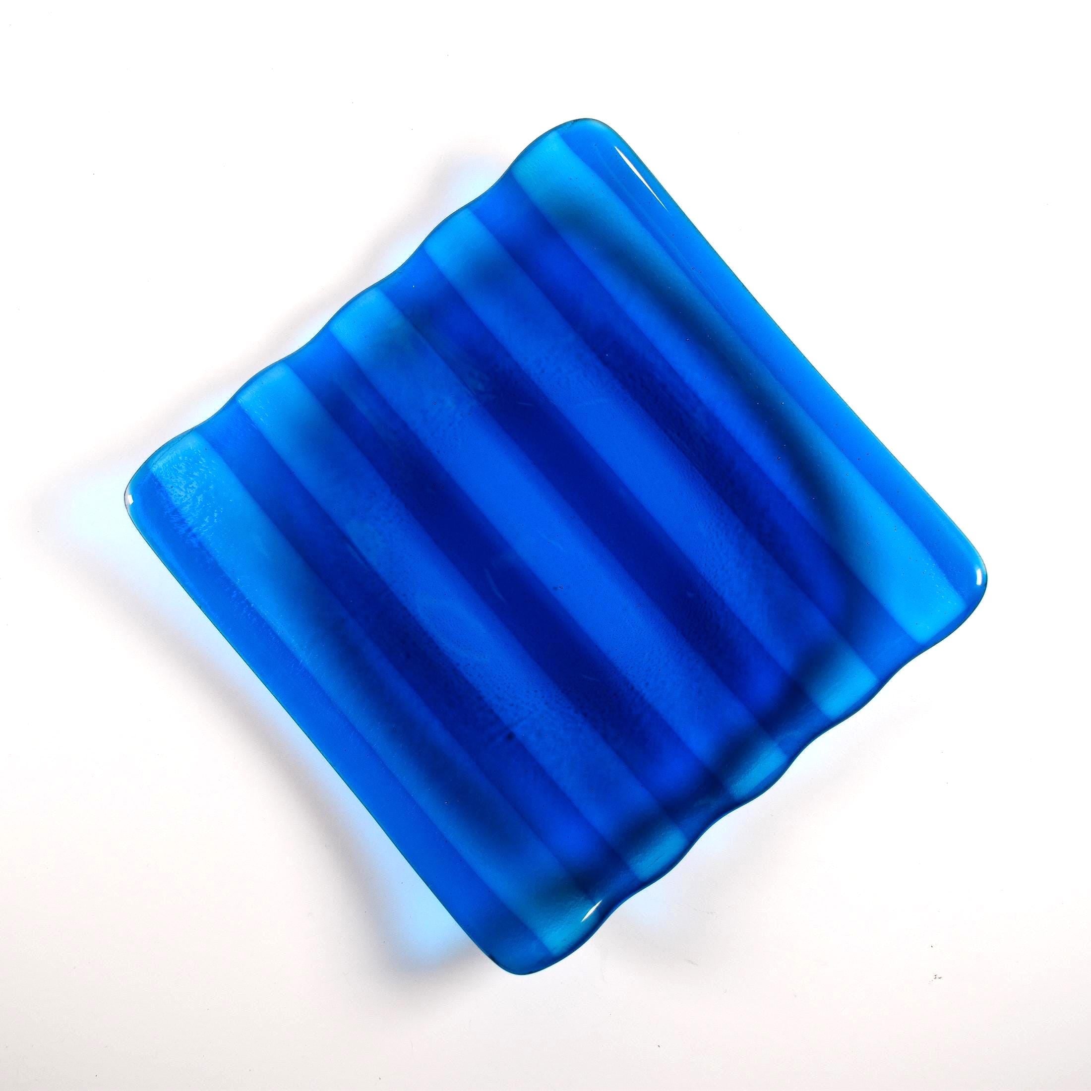 Deep aqua blue fused glass tray with two opposite scalloped edges and strips of a lighter color. The photo is looking down from the top of the tray. The tray is sitting on a white surface.