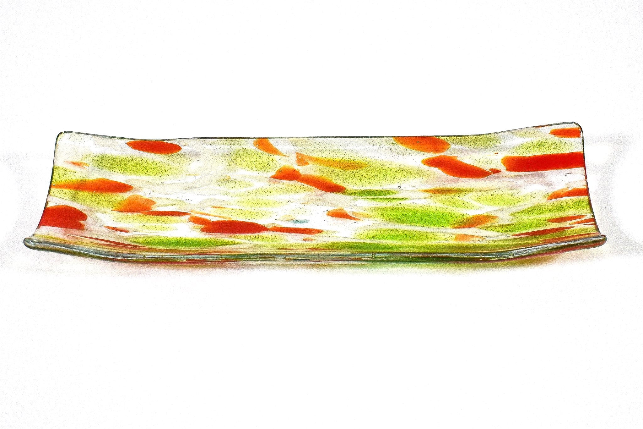 Rectangular glass serving dish with a clear base. The dish features a decorative pattern of orange and green splotches. The colors are distributed across the surface, creating a speckled effect. The edges are slightly curved into a channel shape.