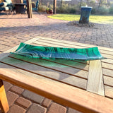 Teal green and white fused glass channel tray with flowing swirls, displayed diagonally on a wooden outdoor table in sunlight.
