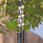 Puffed square silver jasper stone beads, in black, gray and white, are strung with large clear glass round beads, hanging from a tree against a blurred landscape background, anchored by two rectangular pieces of transparent navy blue glass.