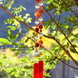 Cinjo Handmade wind chime hanging from tree against foliage background. Two pieces of orange and white glass anchor two legs of beaded wire. Beads include large and small round carnelian beads and large clear glass beads.