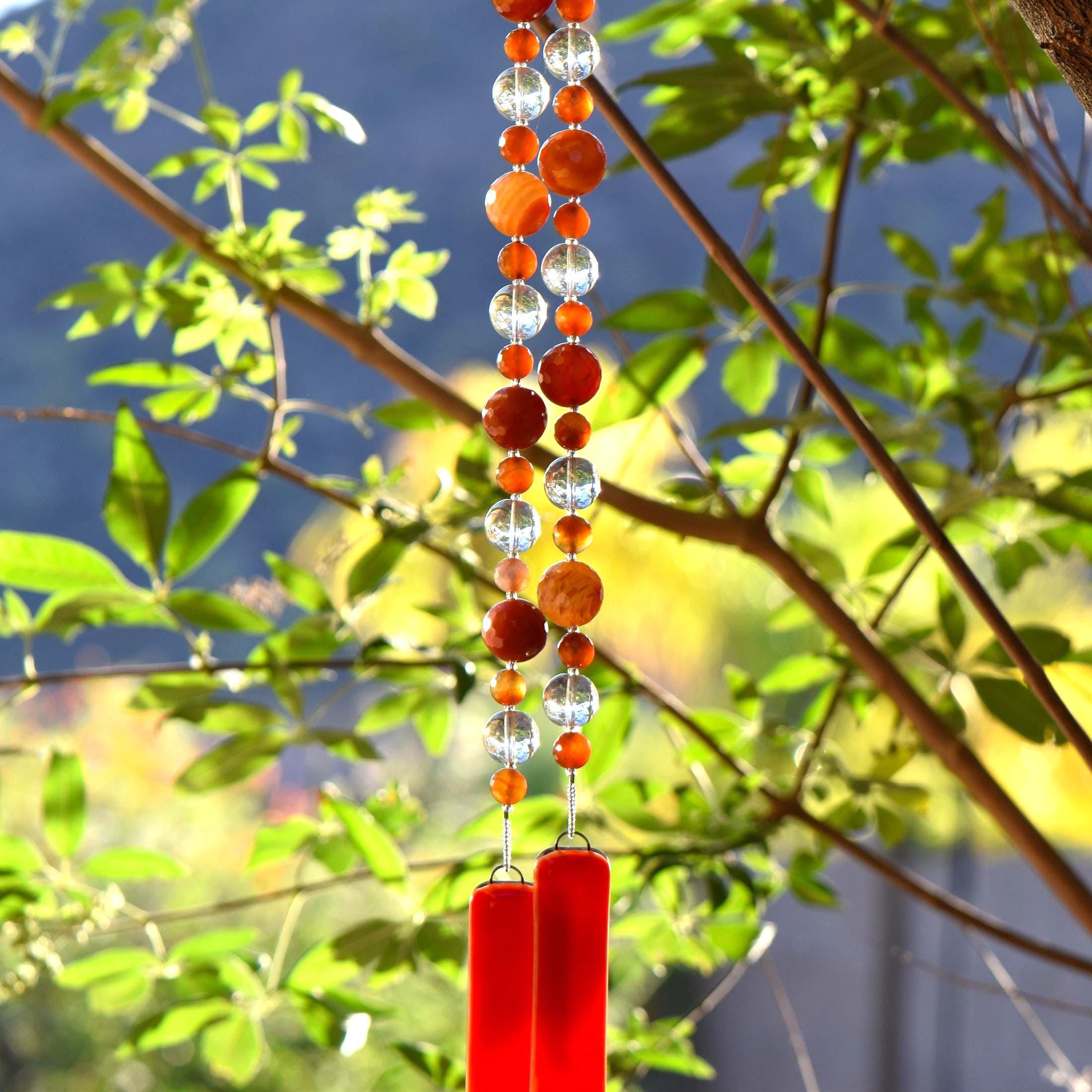 Cinjo Handmade wind chime hanging from tree against foliage background. Two pieces of orange and white glass anchor two legs of beaded wire. Beads include large and small round carnelian beads and large clear glass beads.
