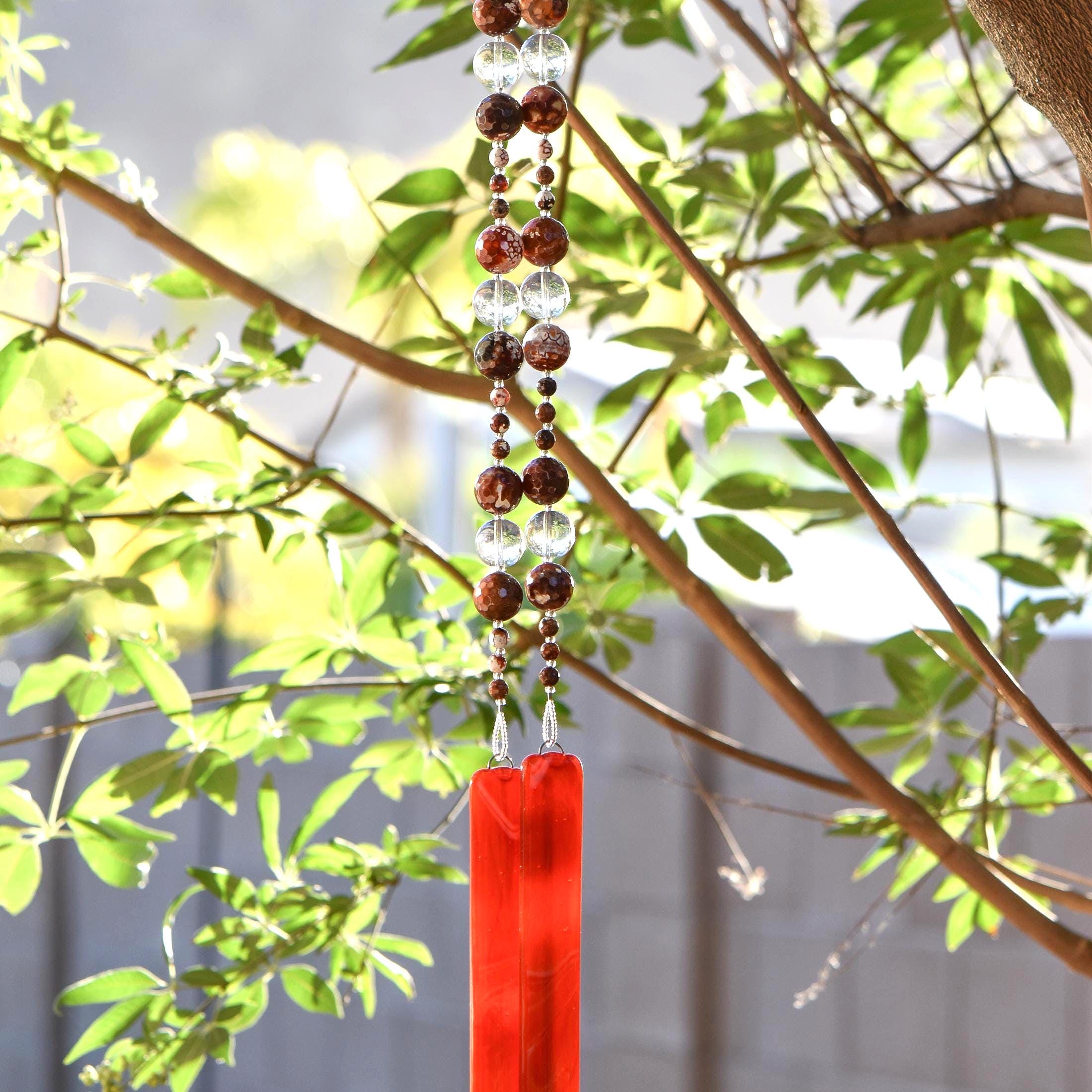 Bottom portion of Cinjo Handmade wind chime hanging from a tree against a background of foliage. The chime is anchored by two rectangular pieces of red and orange fused glass. Above the glass anchors are two strings of beads. The beads are large round carnelian stones, small found carnelian stones and large clear glass beads.