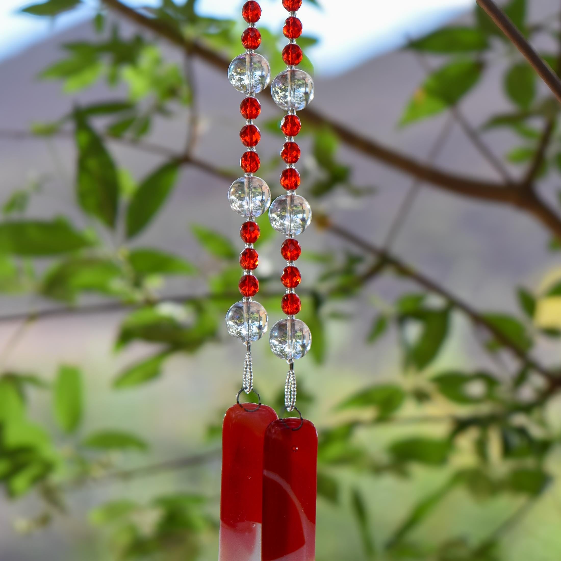 Red and white fused glass wind chime with sparkling red beads and reflective clear glass beads hanging from a tree branch outdoors.