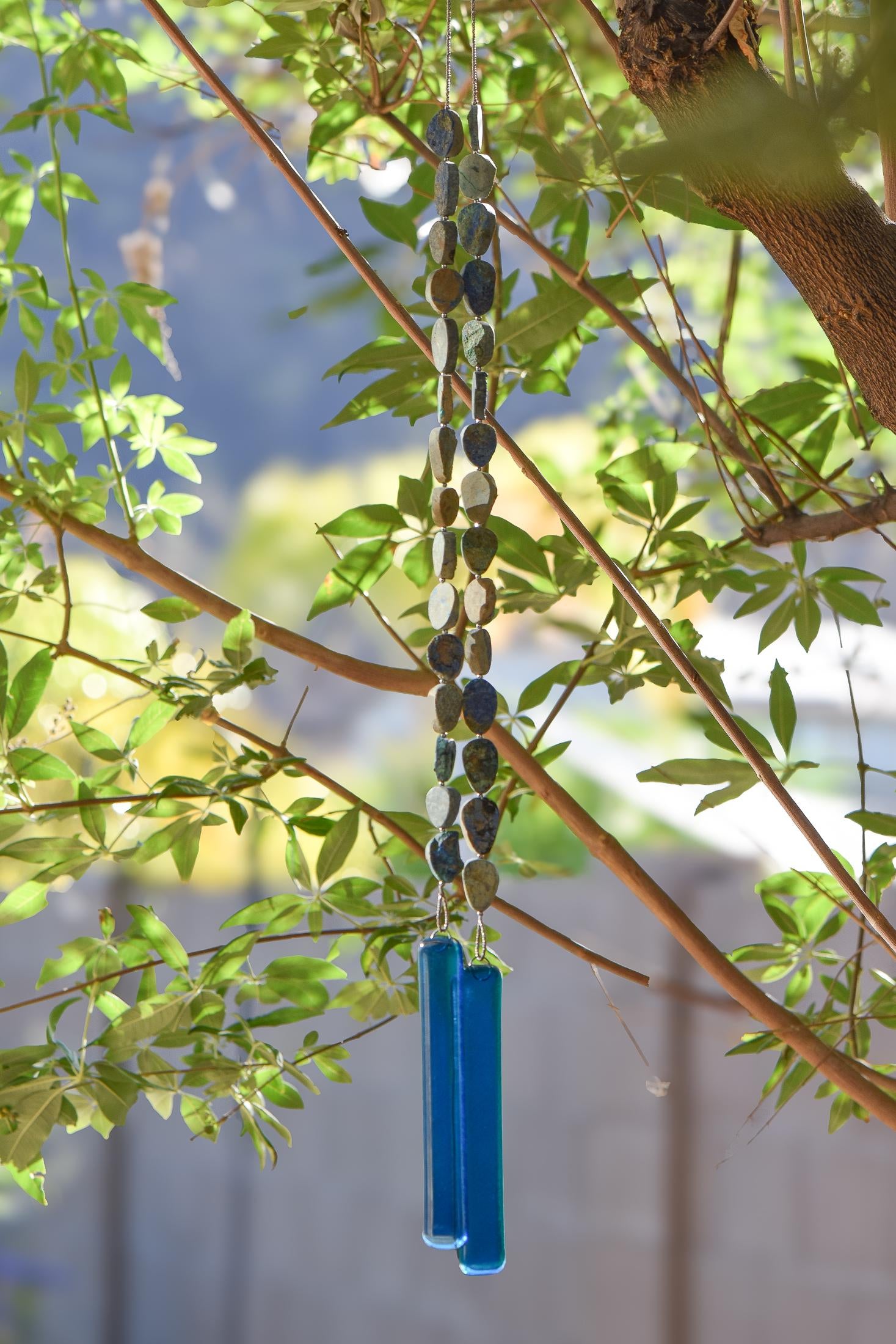 Cinjo Handmade wind chime hanging from tree with foliage in blurred background. Two legs of genuine azurite-malachite stones, roughly cut and with a matte finish are strong on wire and hanging with transparent blue glass pieces at the bottom of each.