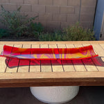 Red and yellow fused glass channel tray with flowing swirled patterns, displayed on a wooden outdoor table in bright sunlight.