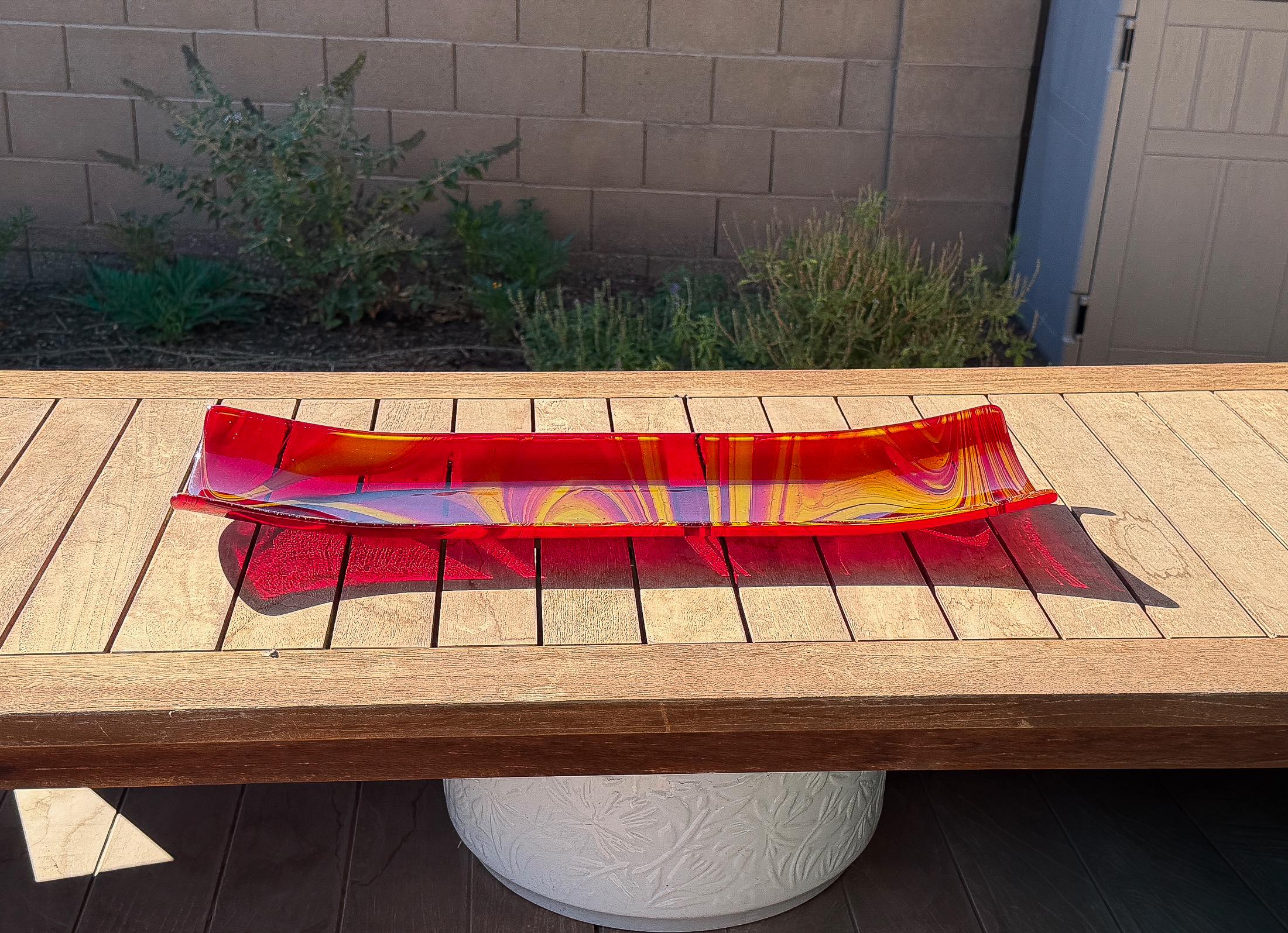 Red and yellow fused glass channel tray with flowing swirled patterns, displayed on a wooden outdoor table in bright sunlight.