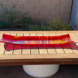 Red and yellow fused glass channel tray with flowing swirled patterns, displayed on a wooden outdoor table in bright sunlight.