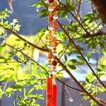 Bottom portion of Cinjo Handmade wind chime hanging from a tree against a background of foliage. The chime is anchored by two rectangular pieces of red and orange fused glass. Above the glass anchors are two strings of beads. The beads are large round carnelian stones, smaller round carnelian stone beads and large clear glass beads.