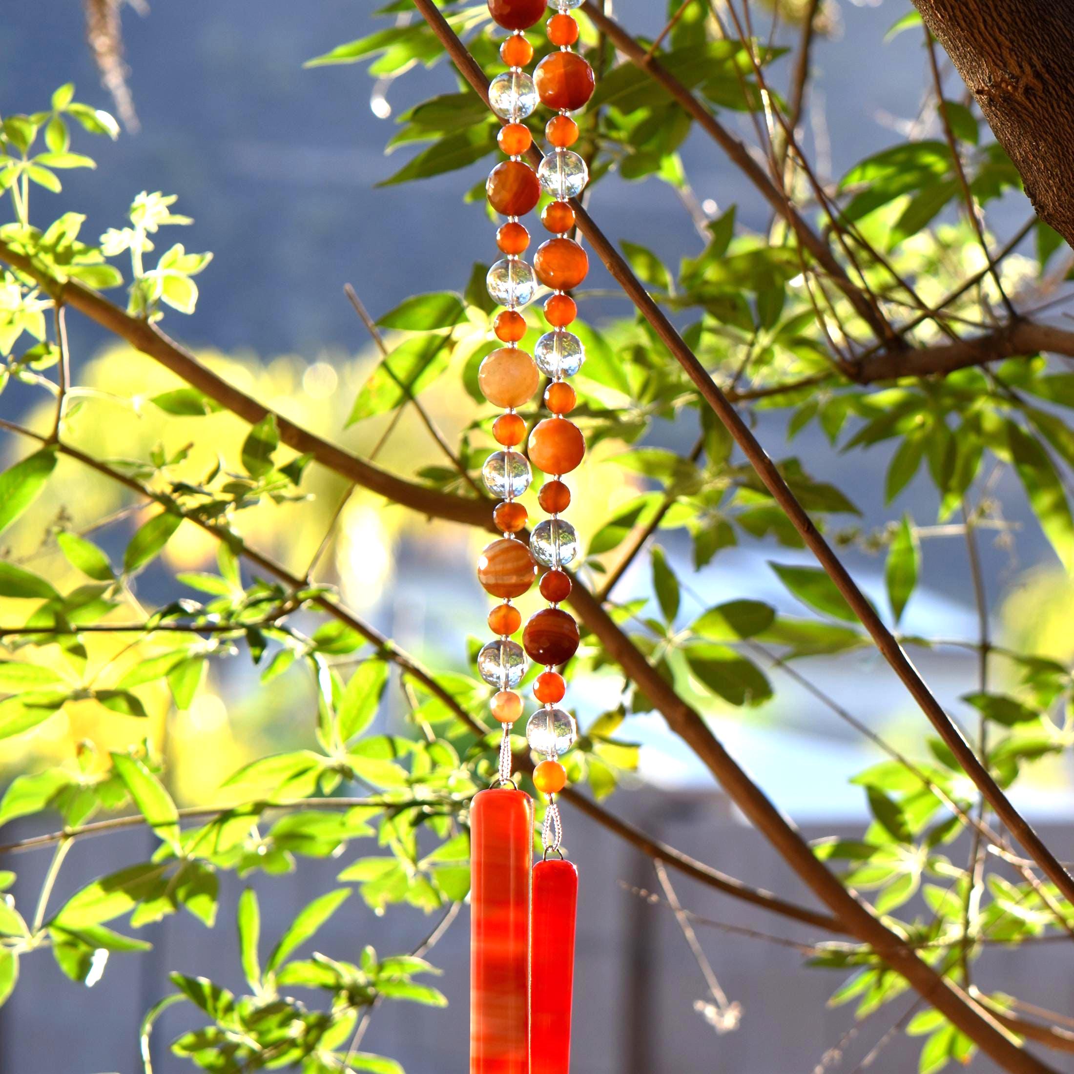 Bottom portion of Cinjo Handmade wind chime hanging from a tree against a background of foliage. The chime is anchored by two rectangular pieces of red and orange fused glass. Above the glass anchors are two strings of beads. The beads are large round carnelian stones, smaller round carnelian stone beads and large clear glass beads.