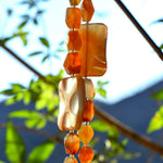 This photo shows the middle section of a Cinjo Handmade wind chime made with two very large slabs of carnelian stone. The two legs of the chime are shown. Each has a single slab of carnelian surrounded by smaller irregular chunks of carnelian strung on stainless steel wire. The chime is hanging from a tree with blurred foliage in the background.