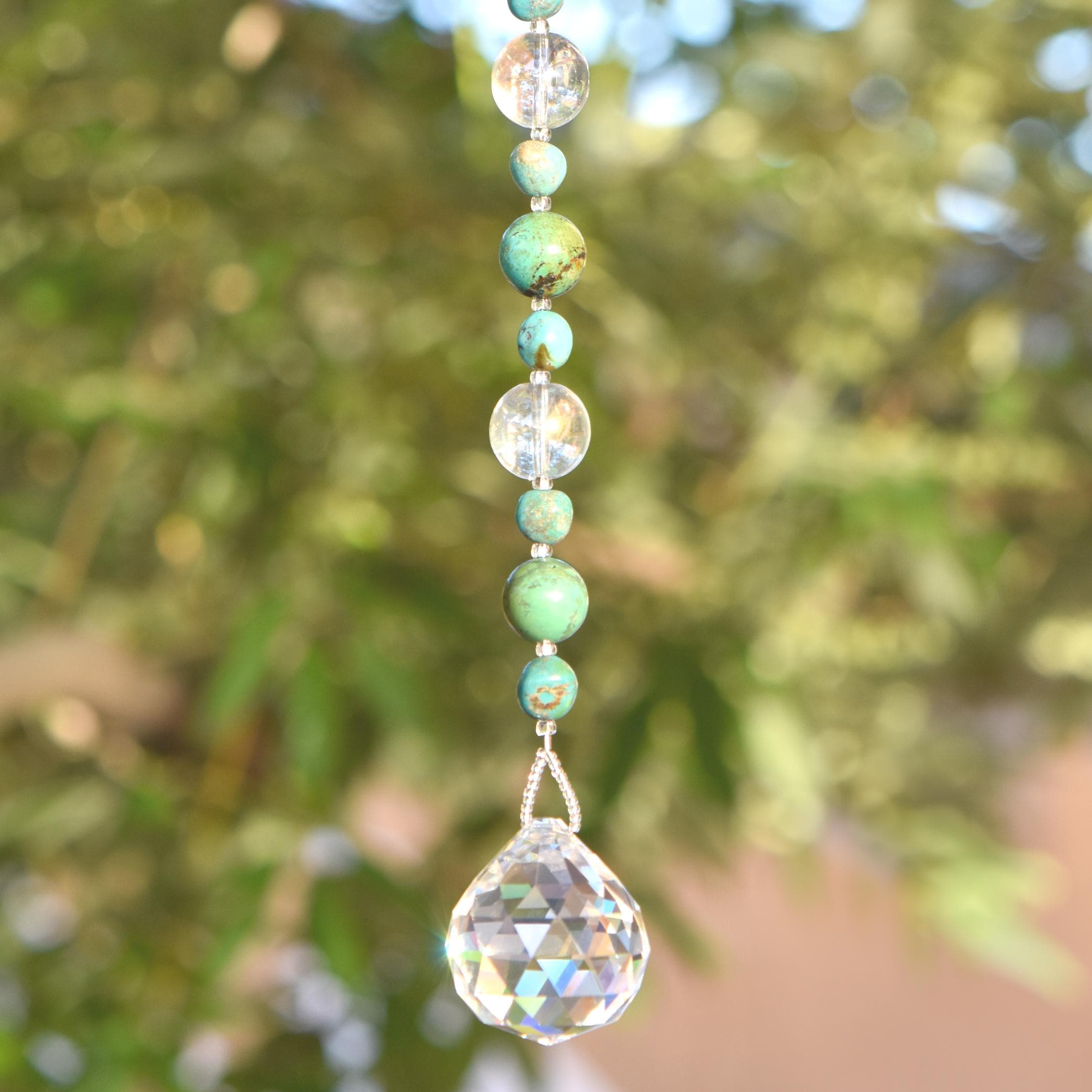 A hanging crystal suncatcher with a string of magnesite stone and clear glass beads. The faceted crystal at the bottom reflects light, creating a sparkling effect. The beads are strung on stainless steel wire that is covered with silver-lined seed beads. Burred foliage background.