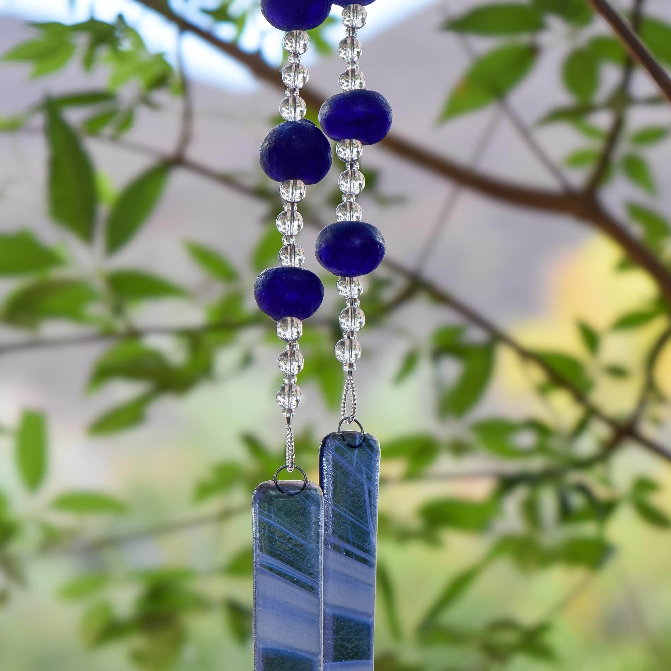 Handmade cobalt blue fused glass wind chime with authentic African Krobo beads and clear glass accent beads hanging outdoors among green leaves.