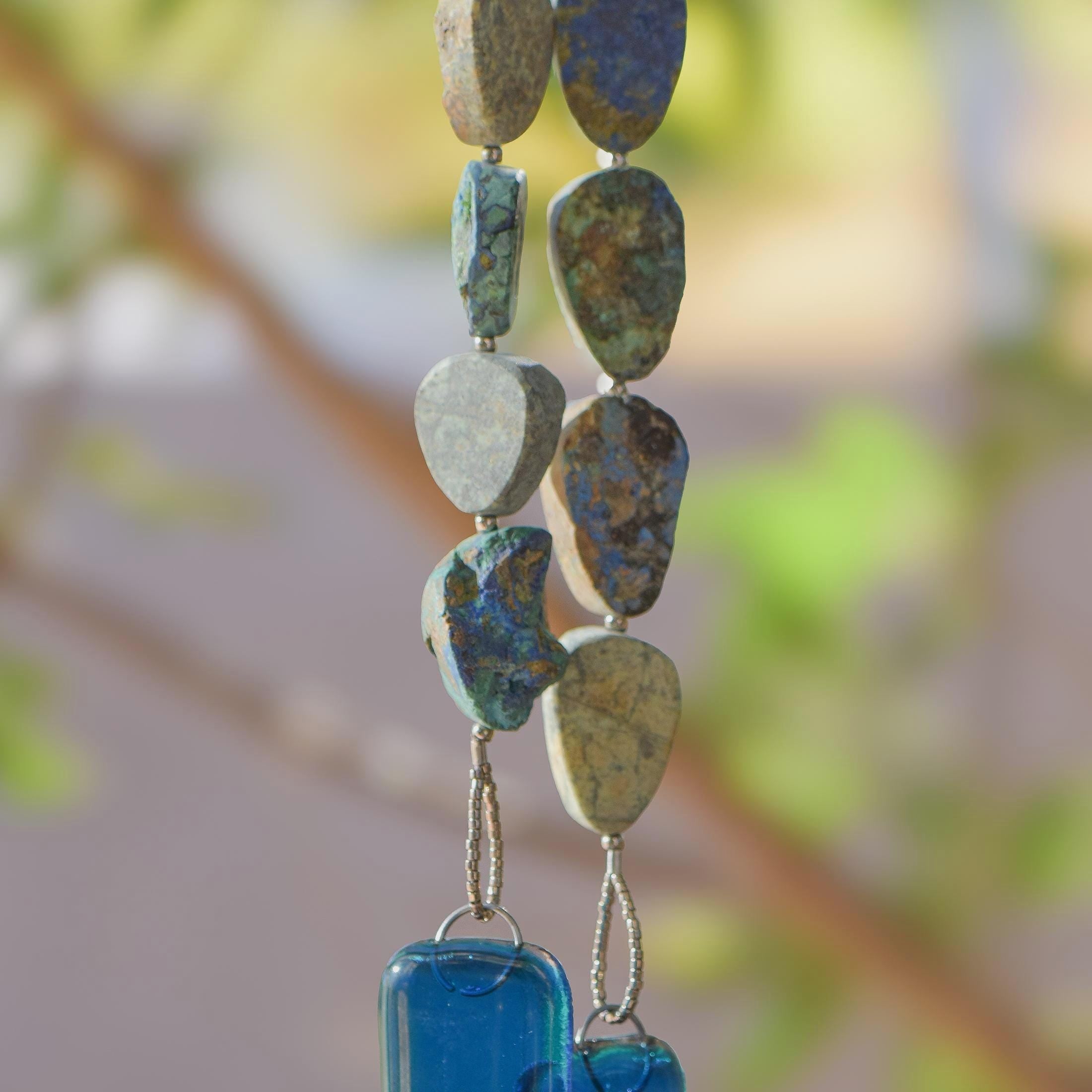 Cinjo Handmade wind chime hanging from tree with foliage in blurred background. Two legs of genuine azurite-malachite stones, roughly cut and with a matte finish are strong on wire and hanging with transparent blue glass pieces at the bottom of each.