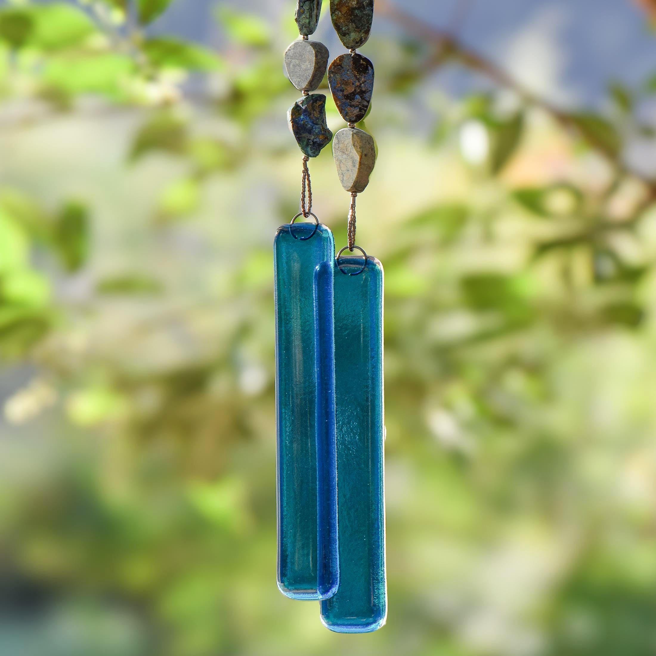 Cinjo Handmade wind chime hanging from tree with foliage in blurred background. Two legs of genuine azurite-malachite stones, roughly cut and with a matte finish are strong on wire and hanging with transparent blue glass pieces at the bottom of each.
