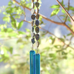Cinjo Handmade wind chime hanging from tree with foliage in blurred background. Two legs of genuine azurite-malachite stones, roughly cut and with a matte finish are strong on wire and hanging with transparent blue glass pieces at the bottom of each.