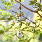 Long handmade sun catcher with star-cut apatite gemstones, copper beads, and a large faceted crystal prism hanging from a tree branch in sunlight.