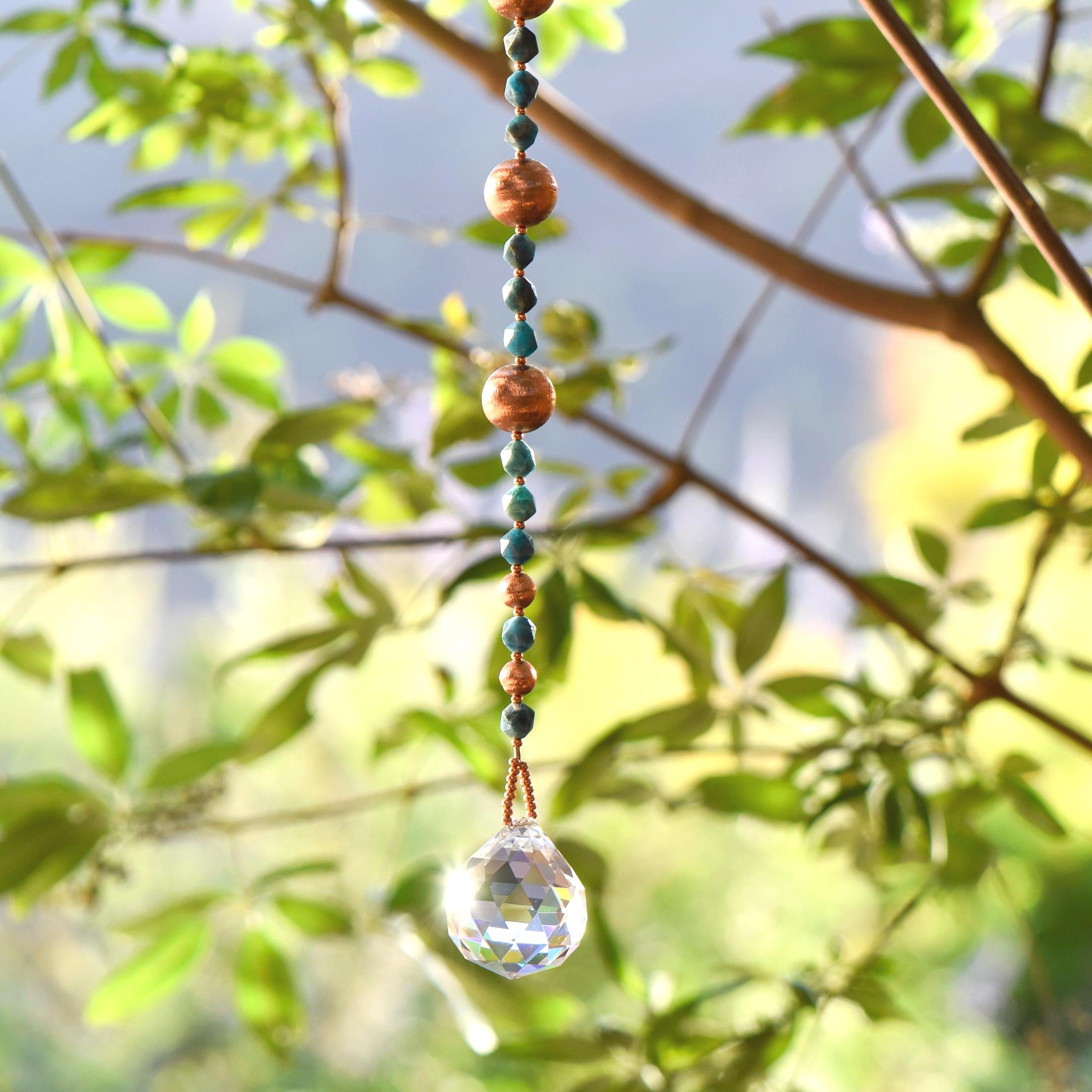 Long handmade sun catcher with star-cut apatite gemstones, copper beads, and a large faceted crystal prism hanging from a tree branch in sunlight.