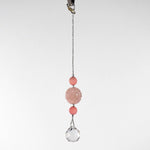Large round carved rose quartz bead flanked by two smaller pink stone beads and anchored by round crystal prism, hanging form a hook in front of white background.