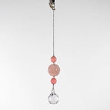 Large round carved rose quartz bead flanked by two smaller pink stone beads and anchored by round crystal prism, hanging form a hook in front of white background.