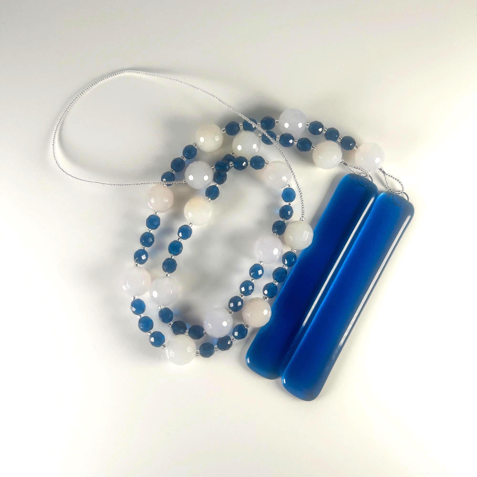 A coiled wind chime with two strings of white and blue beads, and two pieces of dark blue glass, coiled on a gray surface. The beads are interspersed with small blue beads.