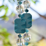 Close up of two beaded strands on a Cinjo Handmade wind chime. Each strand has a large, wavy-rectangular magnesite stone bead flanked by large clear glass beads and smaller blue glass beads.