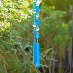 Two beaded wire strings from a Cinjo Handmade chime hanging in front of foliage. Each string has large blue rectangular magnesite stone beads and faceted glass crystal beads, anchored by a piece of turquoise blue fused glass.