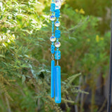 Two beaded wire strings from a Cinjo Handmade chime hanging in front of foliage. Each string has large blue rectangular magnesite stone beads and faceted glass crystal beads, anchored by a piece of turquoise blue fused glass.
