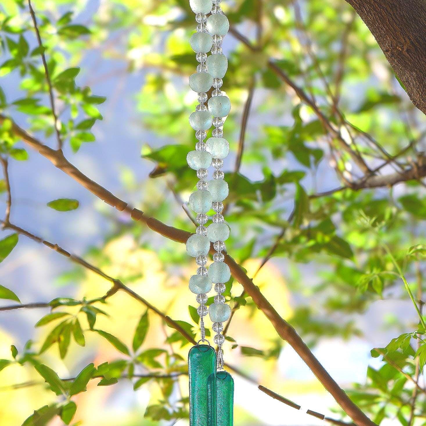 Two strings of beaded stainless steel wire hung in a tree. Each string contains large translucent green Krobo tribe African glass beads paired with smaller clear glass beads, anchored by teal green transparent fused glass.