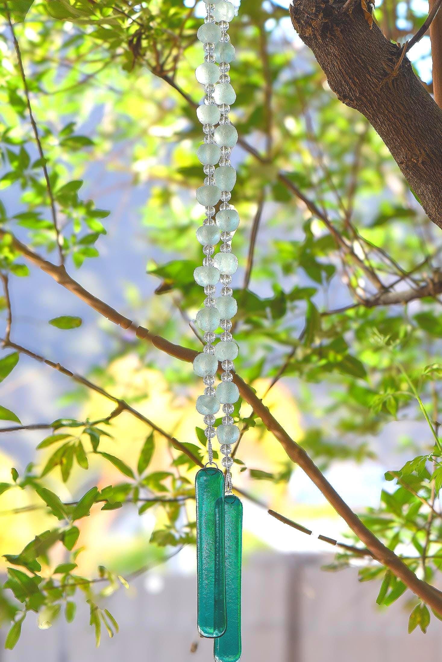 Two strings of beaded stainless steel wire hung in a tree. Each string contains large translucent green Krobo tribe African glass beads paired with smaller clear glass beads, anchored by teal green transparent fused glass.