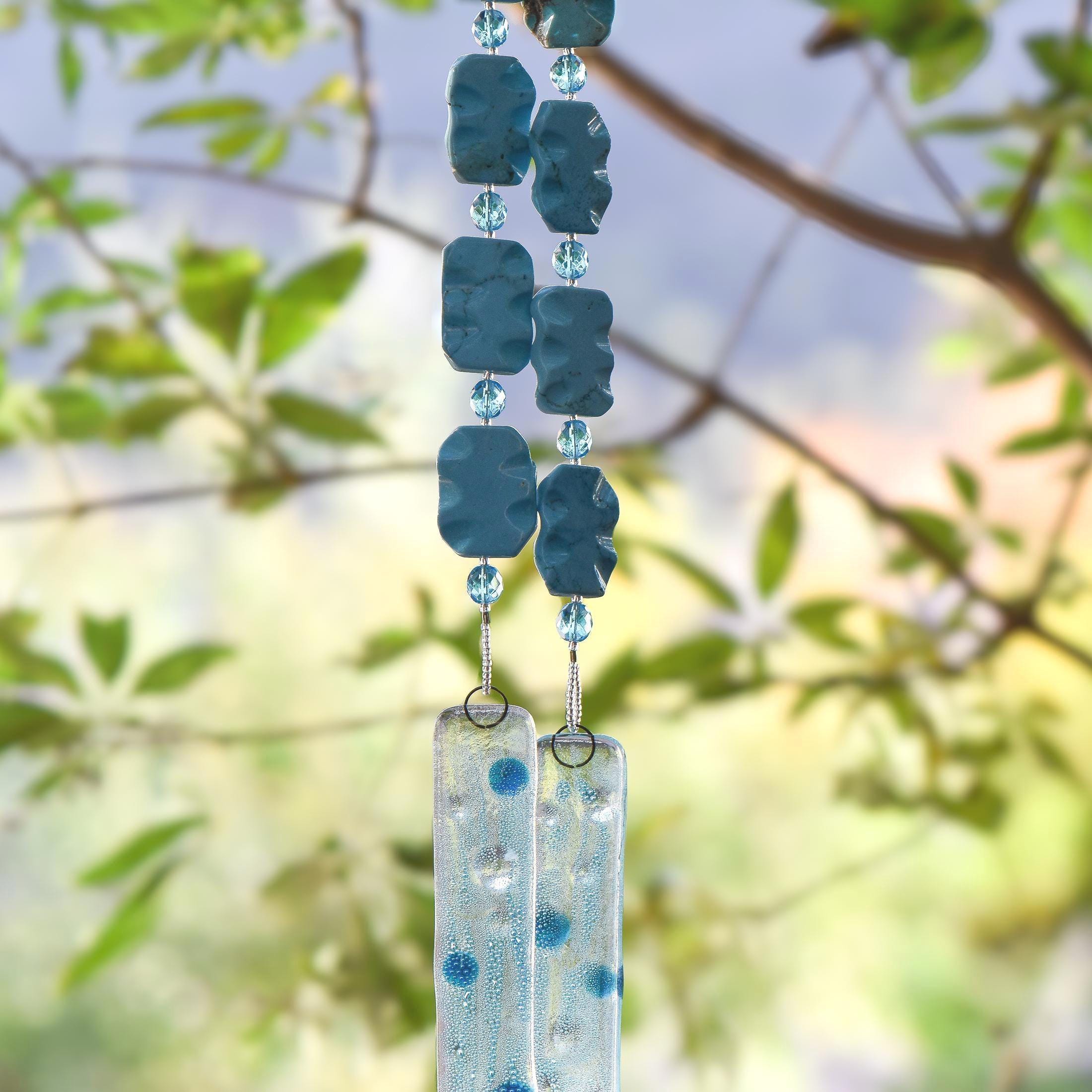 Two strings of stainless steel wire hanging in a tree, covered in large rectangular blue magnesite stone beads, smaller blue glass beads and silver-lined seed beads. Each string is anchored by a single piece of fused glass. The fused glass is clear with added, tiny blue bubbles and accented with large blue and clear dots. Looks like it's underwater. Background is blurred foliage.