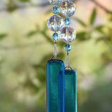 The bottom of two strings of beads, each anchored by a piece of fused transparent blue glass. The strings contain large faceted clear glass crystal beads and smaller transparent blue beads. Against blurred foliage background.